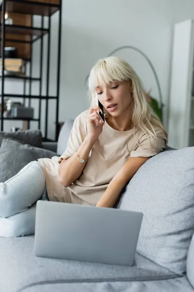 Freelance bionda che parla su smartphone vicino a laptop sfocato sul divano — Foto stock