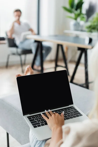 Vista recortada de la mujer escribiendo en el ordenador portátil con pantalla en blanco cerca de novio borroso - foto de stock