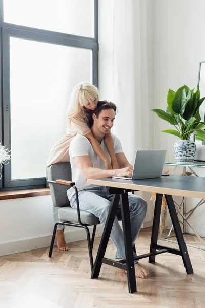 Mujer rubia abrazando novio feliz usando el ordenador portátil mientras trabaja desde casa - foto de stock
