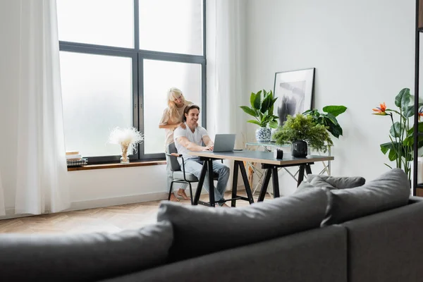 Femme blonde massant les épaules de petit ami travaillant à la maison avec un ordinateur portable — Photo de stock