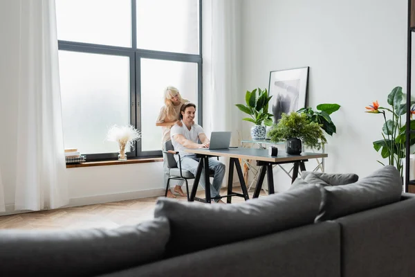 Mujer rubia masajeando hombros de novio trabajando desde casa con laptop — Stock Photo