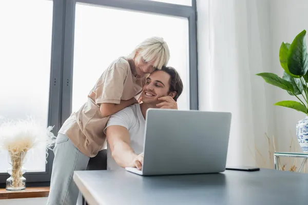 Mujer rubia abrazando novio sonriente usando el ordenador portátil mientras trabaja desde casa - foto de stock