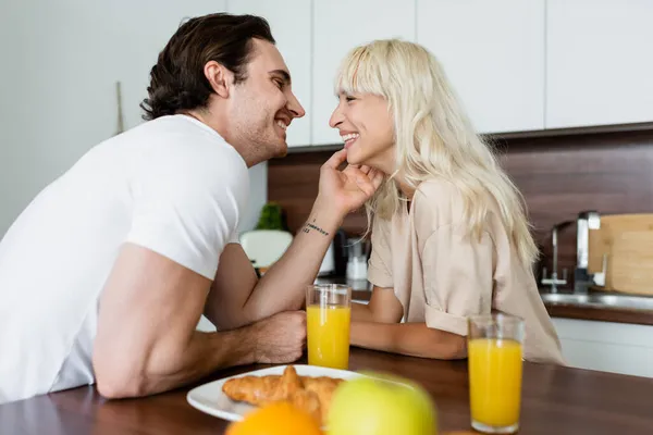 Homem tatuado tocando rosto de namorada feliz perto de óculos com suco de laranja e croissant — Fotografia de Stock