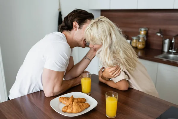 Tattooed man touching face of happy girlfriend near glasses with orange juice and croissants on plate — Stock Photo