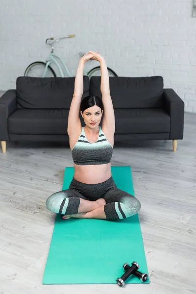 Pregnant woman looking at camera while meditating in lotus pose with raised hands — Stock Photo