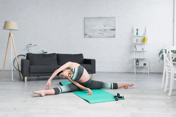 Joven embarazada estiramiento en splits pose en yoga mat en moderno salón - foto de stock