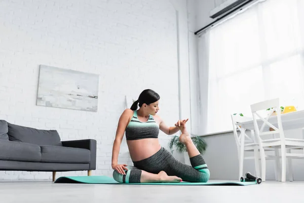 Mujer embarazada estirándose en pose sentada sobre esterilla de yoga en casa - foto de stock