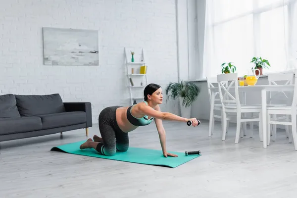 Mujer embarazada joven haciendo ejercicio con la mancuerna mientras está de pie sobre las rodillas en la alfombra de fitness - foto de stock