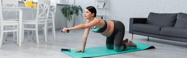 Mujer embarazada de pie sobre las rodillas en la alfombra de fitness, mientras que el entrenamiento con mancuerna en la sala de estar, pancarta - foto de stock