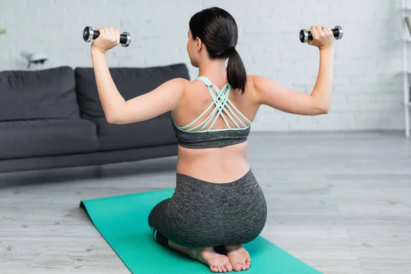 Vista posteriore della donna in abbigliamento sportivo che si allena con i manubri mentre è seduta sul tappeto fitness — Foto stock