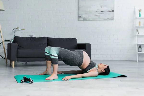 Mujer embarazada en ropa deportiva practicando puente posan sobre esterilla de yoga - foto de stock