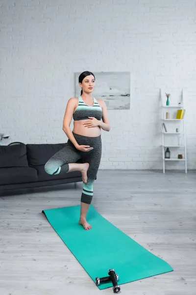 Barfuß schwangere Frau umarmt Bauch beim Meditieren in Baum-Pose zu Hause — Stockfoto