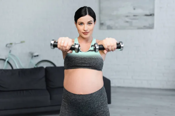 Young pregnant woman looking at camera while training with dumbbells at home — Stock Photo