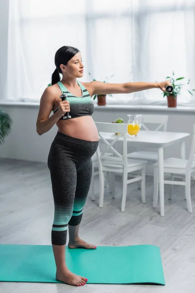 Mujer embarazada descalza entrenando con pesas mientras está de pie en la colchoneta de fitness - foto de stock