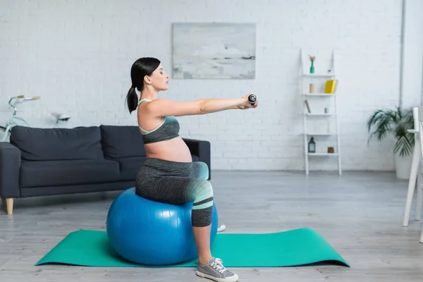 Seitenansicht der brünetten schwangeren Frau, die mit Kurzhanteln trainiert, während sie auf einem Fitnessball sitzt — Stockfoto