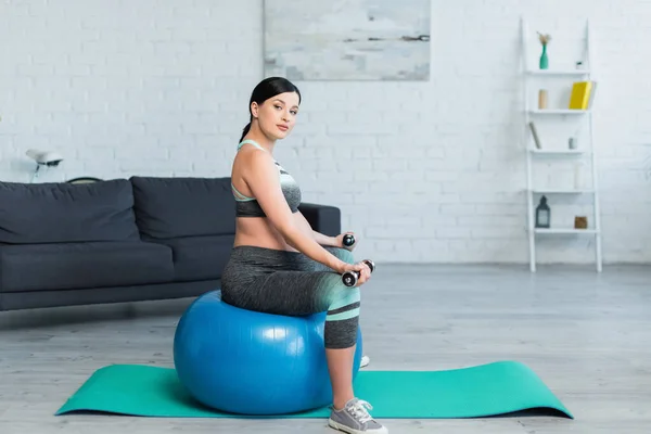 Pretty pregnant woman looking at camera while sitting on fitness ball at home — Stock Photo