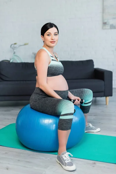 Femme enceinte avec haltères regardant la caméra pendant l'entraînement sur balle de fitness — Photo de stock