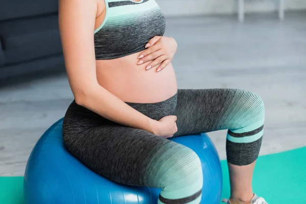 Teilbild einer schwangeren Frau, die ihren Bauch umarmt, während sie auf einem Fitnessball sitzt — Stockfoto