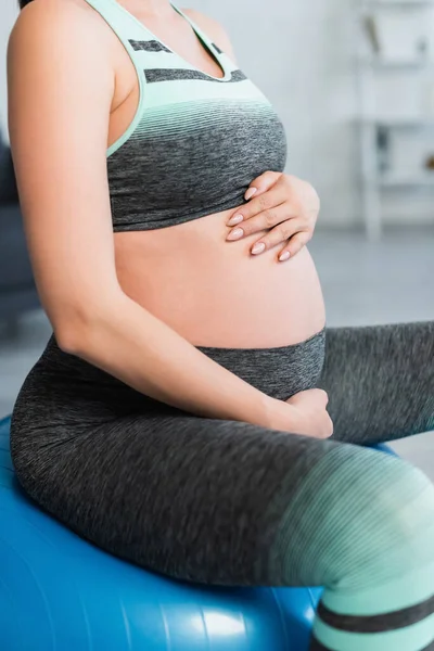 Обрезанный вид беременной женщины в спортивной одежде касаясь живота во время тренировки на фитнес-мяч — стоковое фото