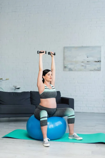 Young pregnant woman with dumbbells in raised hands exercising on fitness ball in living room — Stock Photo