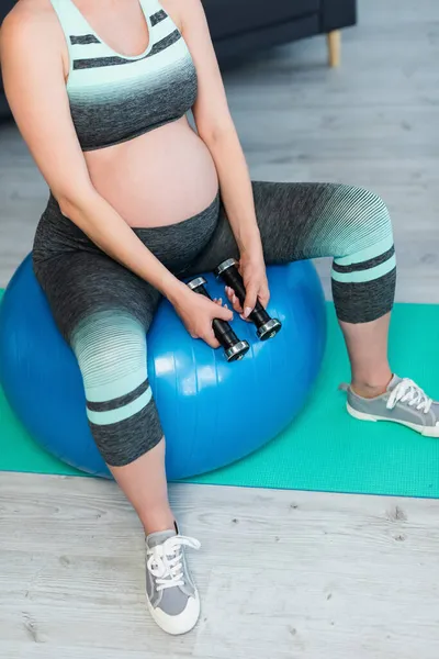 Partial view of pregnant woman holding dumbbells while sitting on fitness ball — Stock Photo