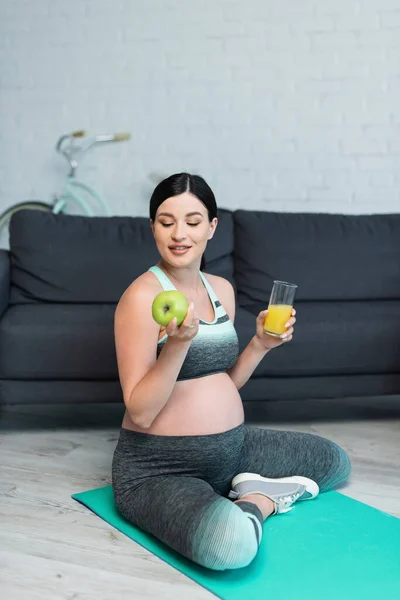 Happy pregnant woman with apple and orange juice sitting with crossed legs on fitness mat — Stock Photo