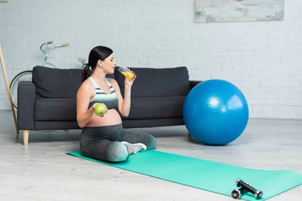 Mujer embarazada sosteniendo manzana y beber jugo de naranja mientras está sentado en la alfombra de fitness - foto de stock
