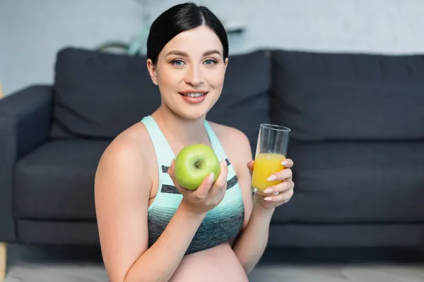 Mujer embarazada joven en sujetador deportivo sonriendo a la cámara mientras sostiene la manzana y el jugo de naranja - foto de stock