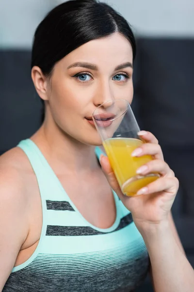 Mujer joven en sujetador deportivo mirando a la cámara mientras bebe jugo de naranja fresco - foto de stock