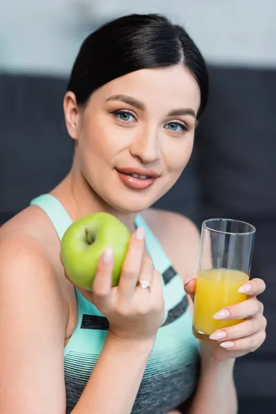 Pretty, brunette woman holding ripe apple and fresh orange juice while smiling at camera — Stock Photo