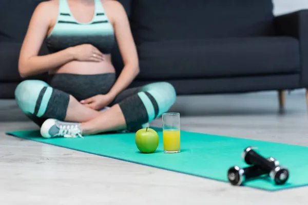 Обрезанный вид размытой беременной женщины медитирующей в позе лотоса рядом со свежим яблоком, апельсиновым соком и гантелями — стоковое фото