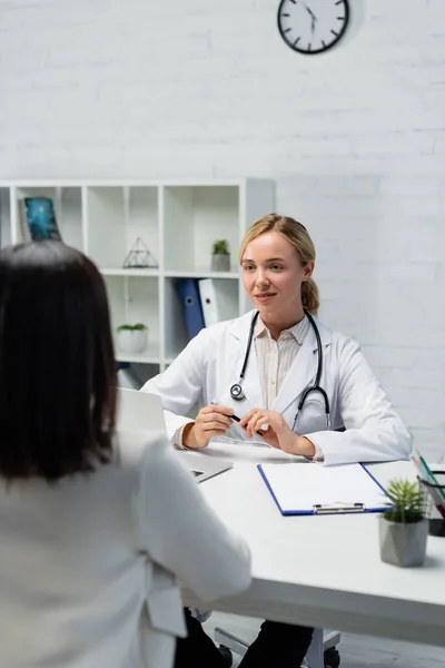 Jeune médecin assis près d'un ordinateur portable et presse-papiers vide lors d'un rendez-vous avec une femme brune au premier plan flou — Photo de stock