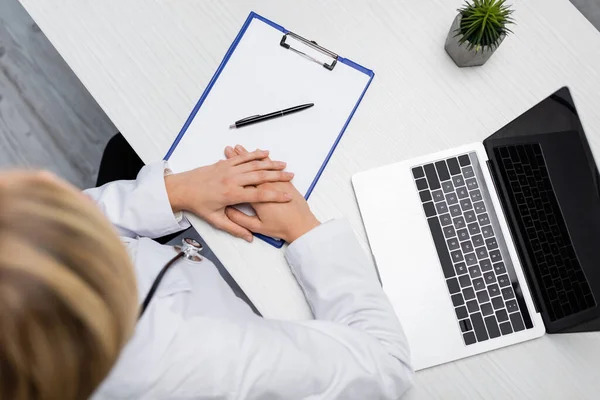 Draufsicht des verschwommenen Arztes am Schreibtisch neben Laptop und Stift auf leerem Klemmbrett — Stockfoto