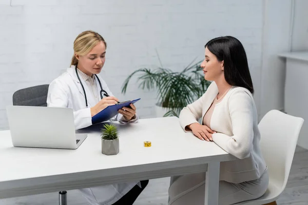 Doctor escribiendo en portapapeles cerca de la mujer embarazada y portátil en el escritorio - foto de stock