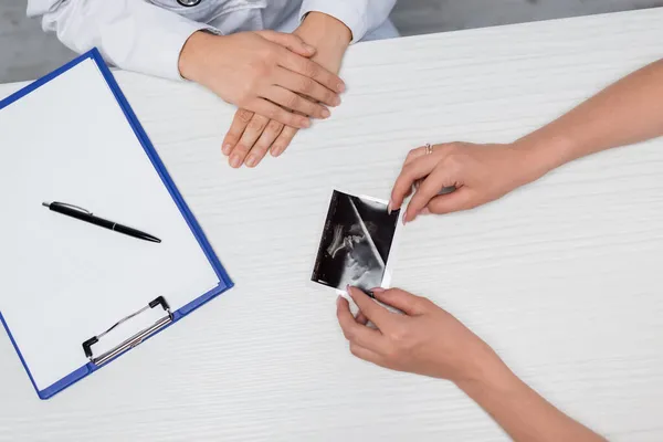 Partial view of doctor and woman near ultrasound scan with pregnancy confirmation and blank clipboard on desk — Stock Photo