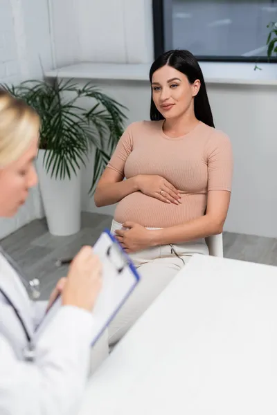 Glücklich schwangere Frau sieht verschwommenen Arzt, der Rezept auf Klemmbrett schreibt — Stockfoto