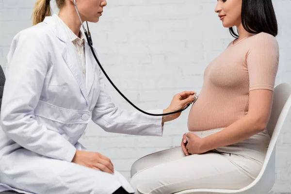 Partial view of pregnant woman and doctor examining her belly with stethoscope during consultation — Stock Photo