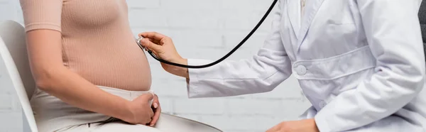 Частичный взгляд врача, осматривающего живот беременной женщины стетоскопом, баннер — стоковое фото