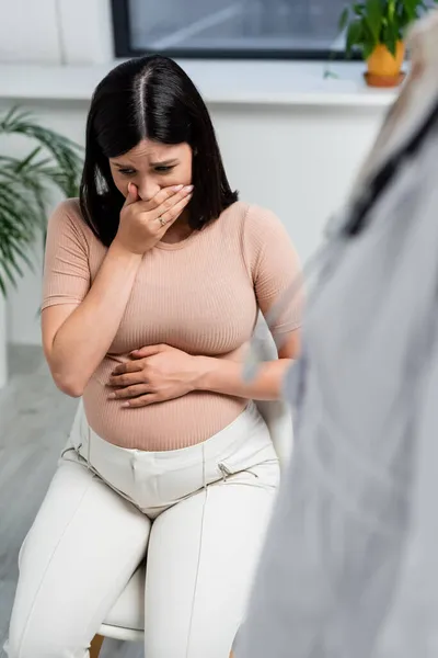 Upset pregnant woman covering mouth with hand near blurred doctor in consulting room — Stock Photo