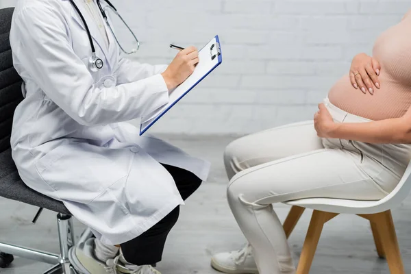 Частковий вигляд вагітної жінки, що сидить поруч з лікарем, пишучи на кишені — стокове фото