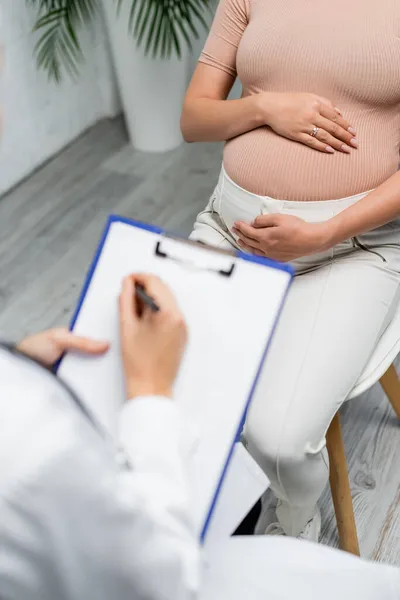 Cropped view of blurred doctor writing on clipboard during appointment with pregnant woman — Stock Photo