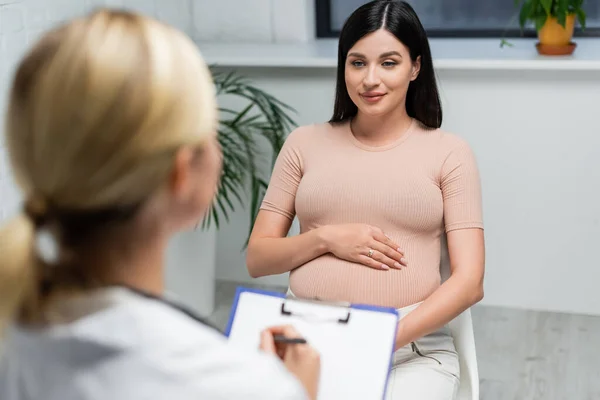 Беременная женщина рядом с размытым врачом выписывает рецепты в консультационной комнате — стоковое фото