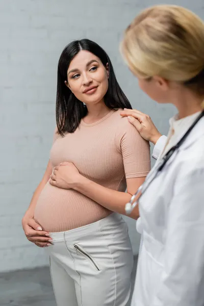 Mulher grávida sorrindo enquanto médico borrado tocando seu ombro durante a consulta — Fotografia de Stock