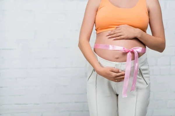 Partial view of pregnant woman with satin ribbon on belly standing near white wall — Stock Photo
