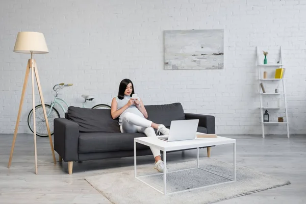 Mulher grávida usando smartphone enquanto se senta no sofá na sala de estar perto do laptop na mesa — Fotografia de Stock