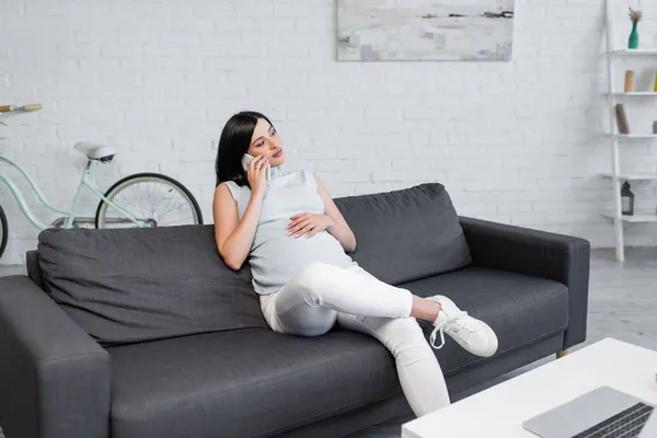 Young pregnant woman talking on smartphone in living room near blurred laptop on table — Stock Photo