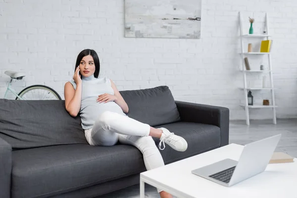 Brunette pregnant woman calling on mobile phone while sitting on sofa near blurred laptop on table — Stock Photo