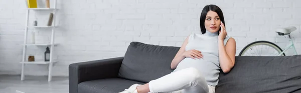 Brunette pregnant woman talking on mobile phone on couch in living room, banner — Stock Photo