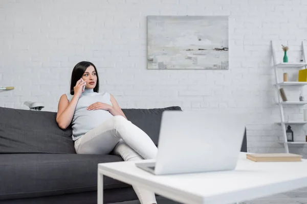 Joven mujer embarazada hablando en el teléfono inteligente en el sofá cerca borrosa portátil en la mesa - foto de stock