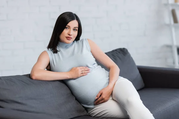 Jeune femme enceinte embrassant le ventre tout en étant assis sur le canapé à la maison — Photo de stock